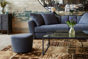  Polly Ottoman in Bellamy Slate next to a glass coffee table and a shelby sofa. Photographed in Bellamy Slate. 