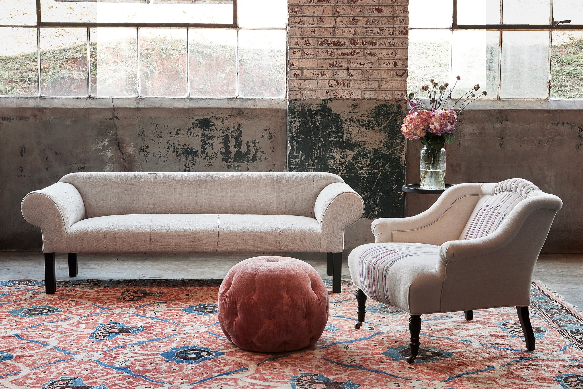  Sofa and chair in front of large windows with a pink velvet pouf on a colorful rug. Photographed in Velluto Rose. 