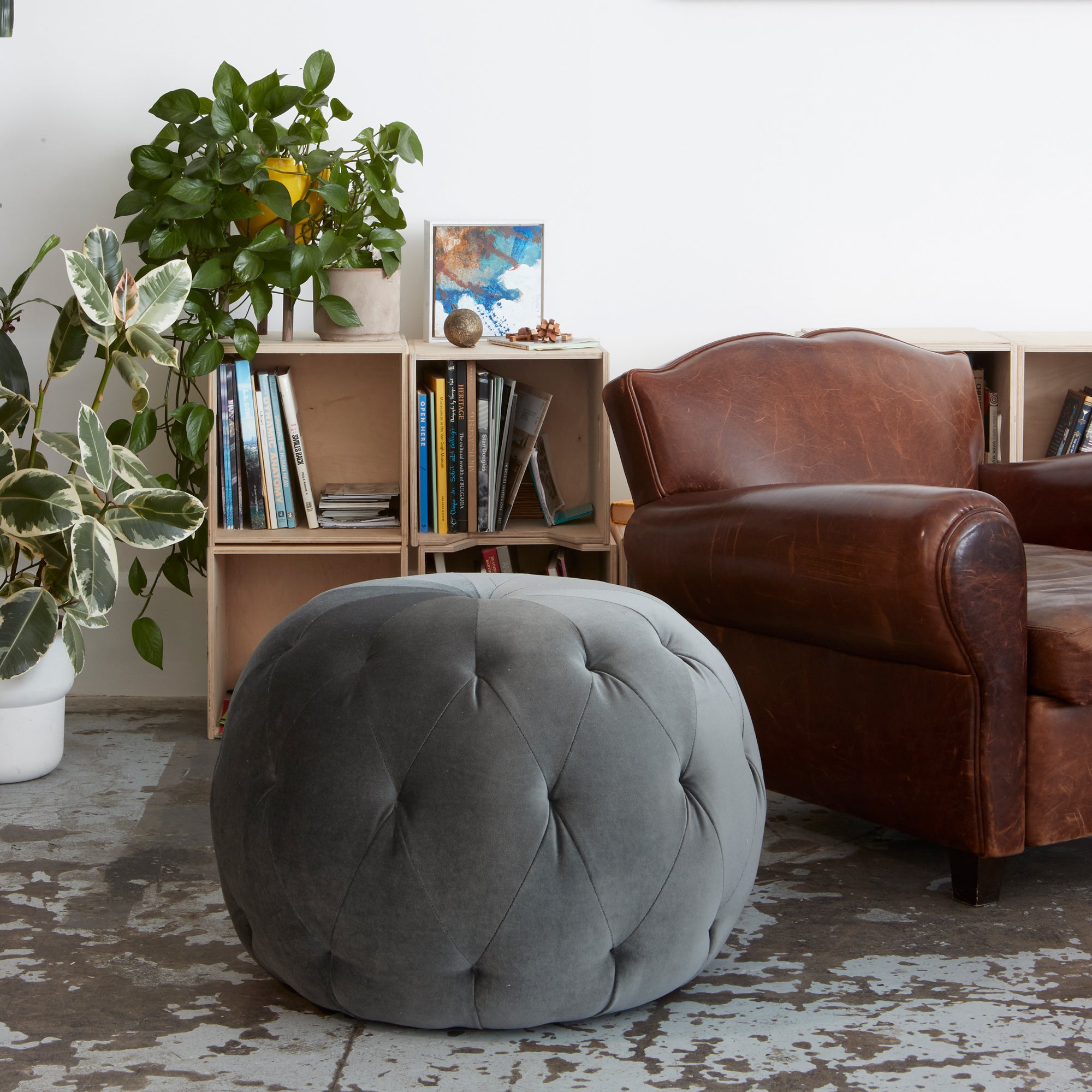  Pouf Ottoman 26 in Matteo Dark Grey next to a leather chair and small bookcase. Photographed in Matteo Dark Grey. 