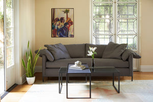  The Radley is upholstered in Logan Grey, photographed in a living room with plenty of daylight coming through a large door window behind the sofa. A painting representing 4 people is on the back wall. The coffee table in front is made with 2 rectangular coffee table on top of each other, one has a metal top and the other a mirror top.There is a vase with white flowers and some books on top.There is a snake plant in a white pot on the floor, to the left of the sofa. Photographed in Logan Grey. 