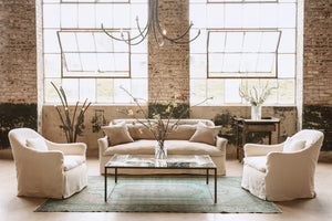  Slipcovered sofa with matching set of chairs in light neutral fabric with rectangular coffee table between them all and a ramo chandelier hanging above.  
