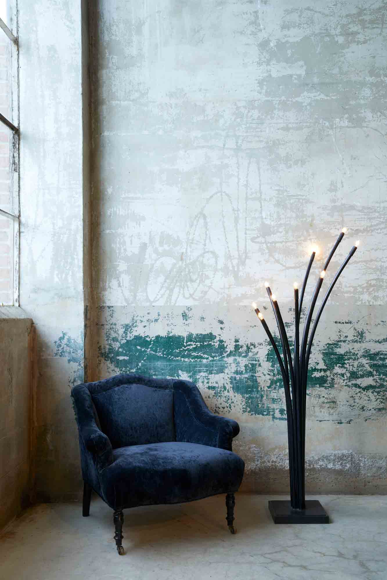  Upholstered chair in dark blue fabric with ramo floor lamp next to it , both placed against distressed brick wall and large window.  