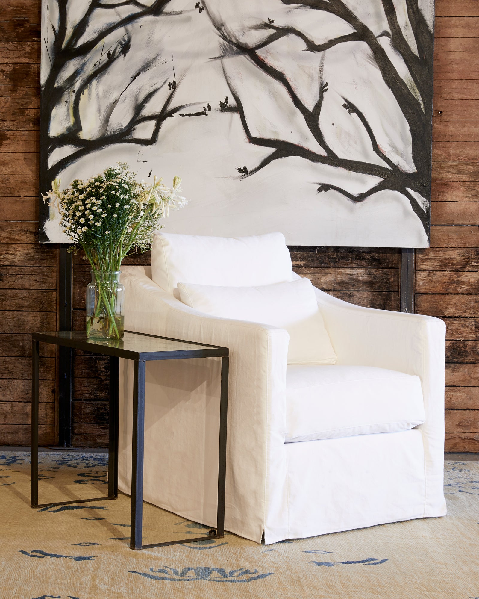  Rebecca chair in Denim White next to a metal side table. In the background is wood wall with a large painting of tree branches. Photographed in Denim White. 