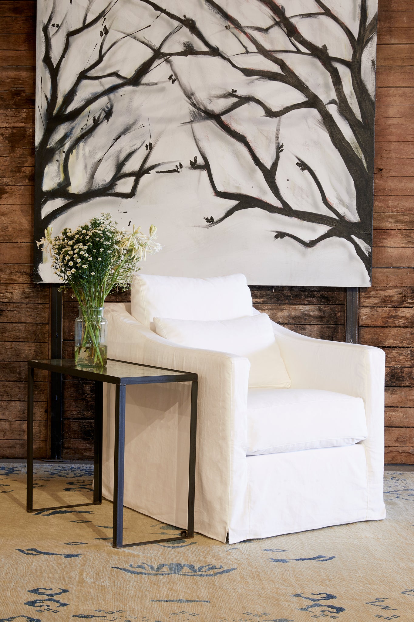  Rebecca chair in Denim White next to a metal side table. In the background is a wood wall with a painting of tree branches. Photographed in Denim White. 