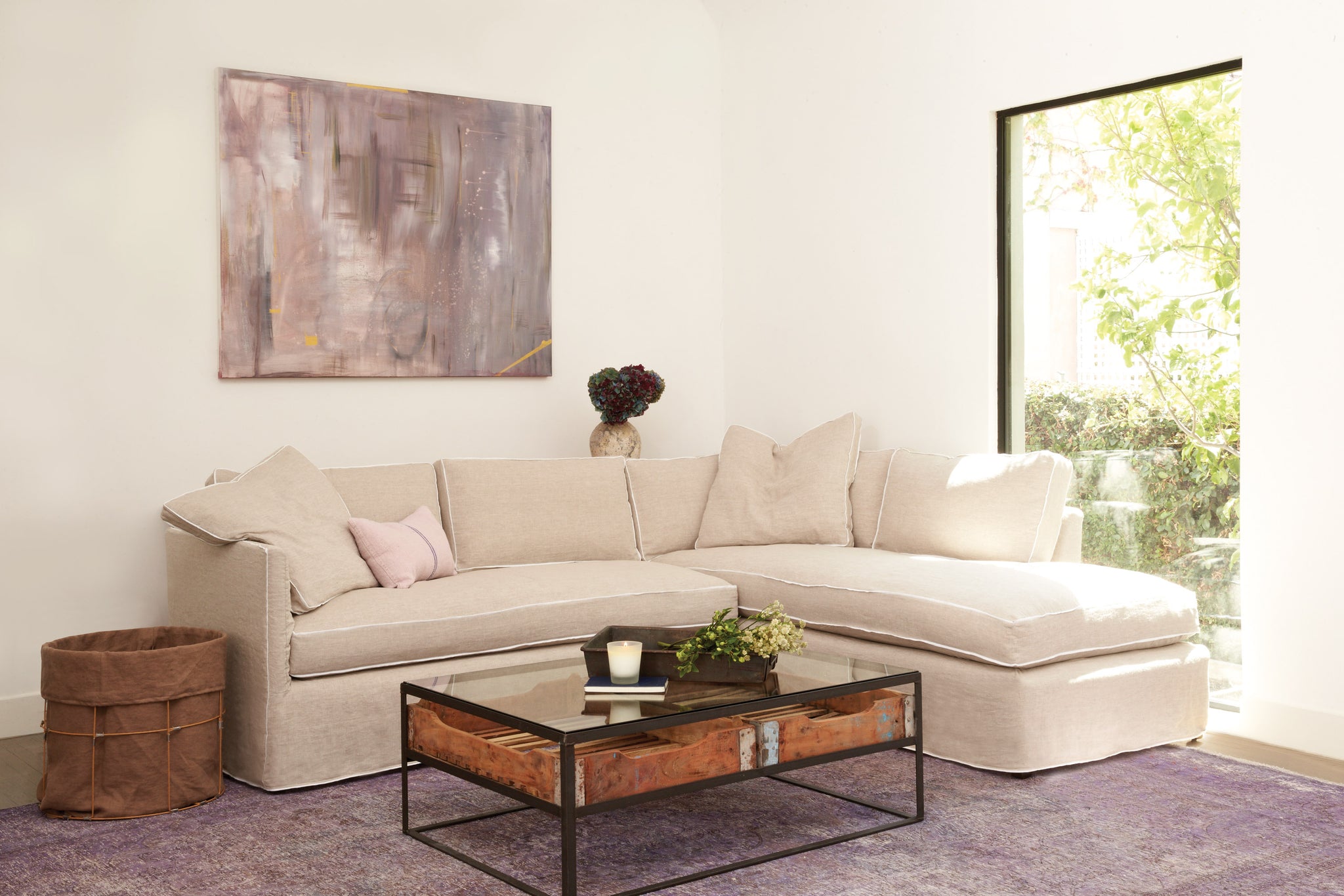 Renata Sectional in Logan Oatmeal with Logan White Flat Welt next to a glass coffee table. IN the background we see a single painting on the wall. Underneath the sectional is a dark colored rug. Photographed in Logan Oatmeal. 