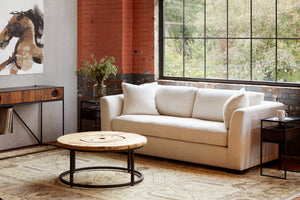 Ryder sofa in Lester Snow in a loft style room. Round coffee table with wood top in front. In the background, large window, and wood and metal side table on each side with a vase with greenery on the left. A wood console is on the left of the picture with a painting of a horse above it. Photographed in Lester Snow. 
