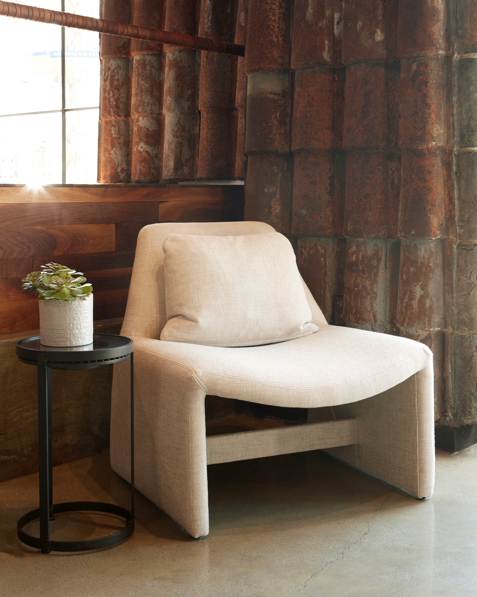  Chair in front of a wall with brown tiles. A round side table on the left with a plant on top. Photographed in Bellamy Natural. 