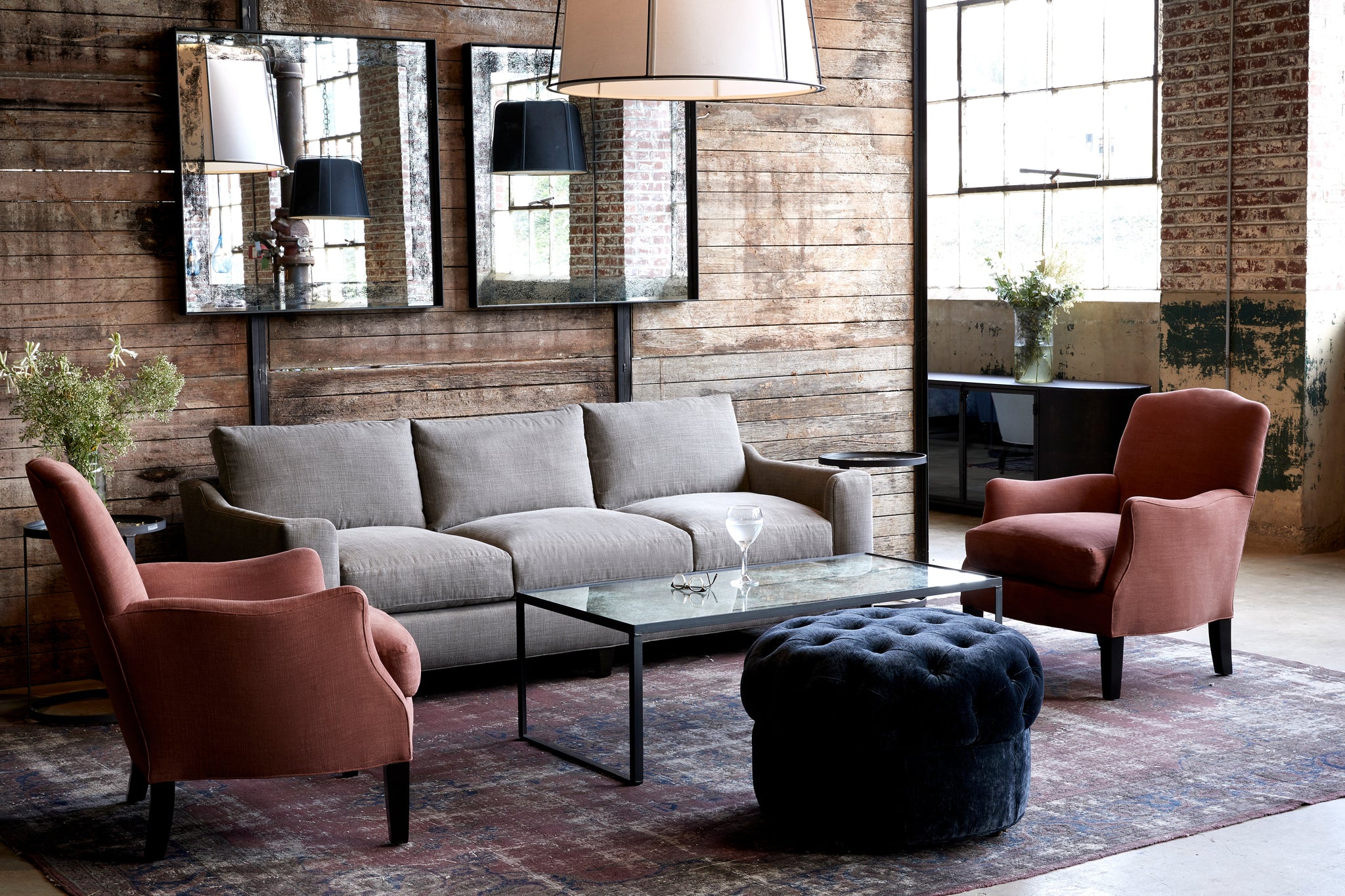  The Gunner sofa is in Rye Warm Grey in a showroom with a wood wall in the back, 2 square mirrors hanging. There are 2 Sebastian chairs in a red linen on the sides, a metal coffee table with a glass top in the middle and a round tufted ottoman in black velvet in front. 