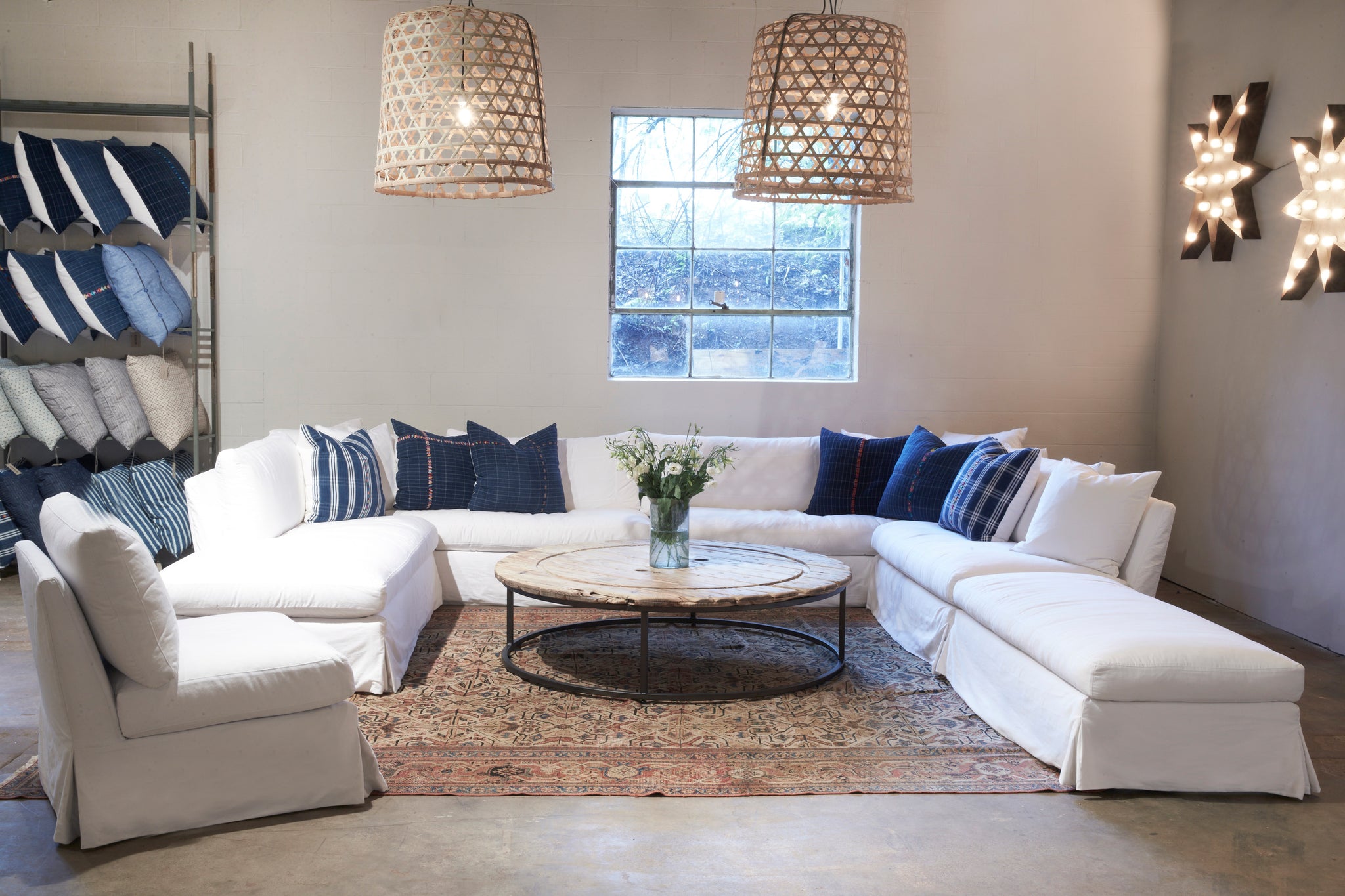  Seda bench in Denim White next to a large white sofa and chair. In the center is a wood coffee table. Above all the pieces hangs two chandeliers. You can also see multiple pillows on a rack in the background. Photographed in Denim White. 