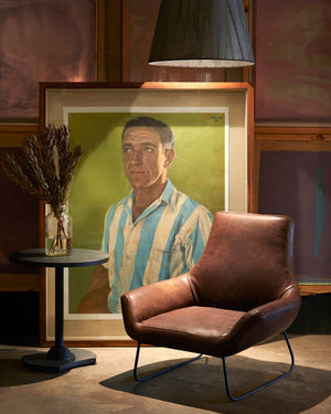  Brown leather chair in front of a large painting of a man in a stripped shirt. A round metal side table is on the left with a vase and flowers. Photographed in Spur Chocolate. 
