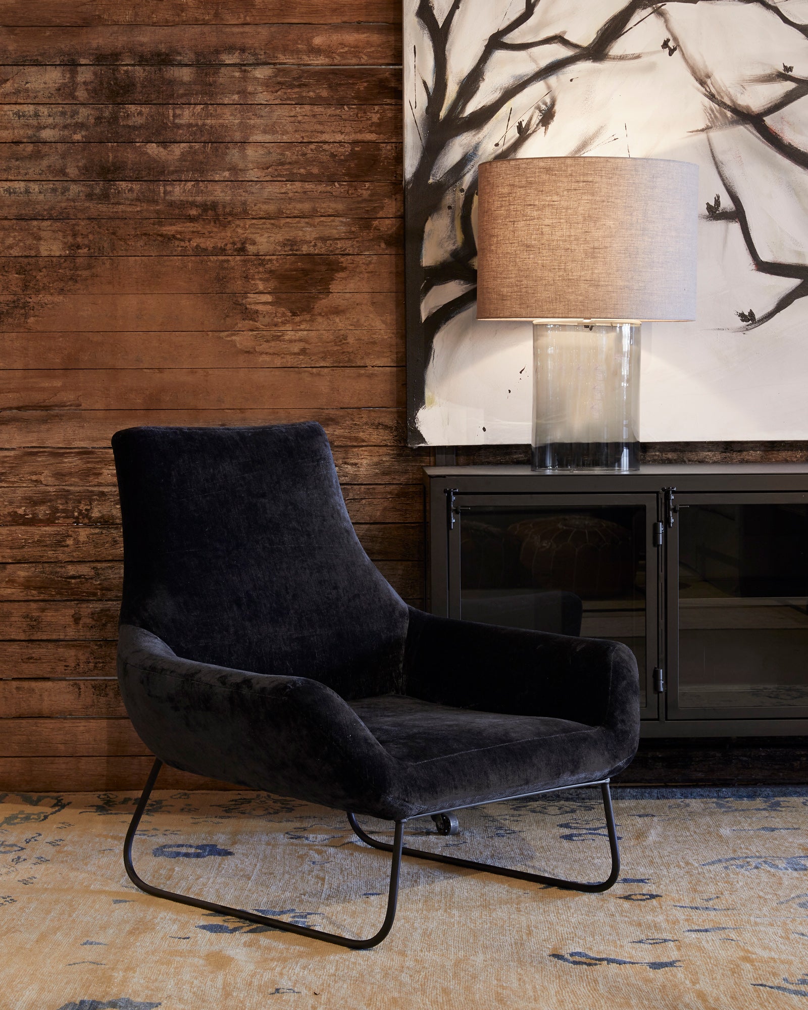  Black velvet chair in front of a wood wall and a metal console with a Cylinder Table Lamp on top. Photographed in Velluto Midnight. 
