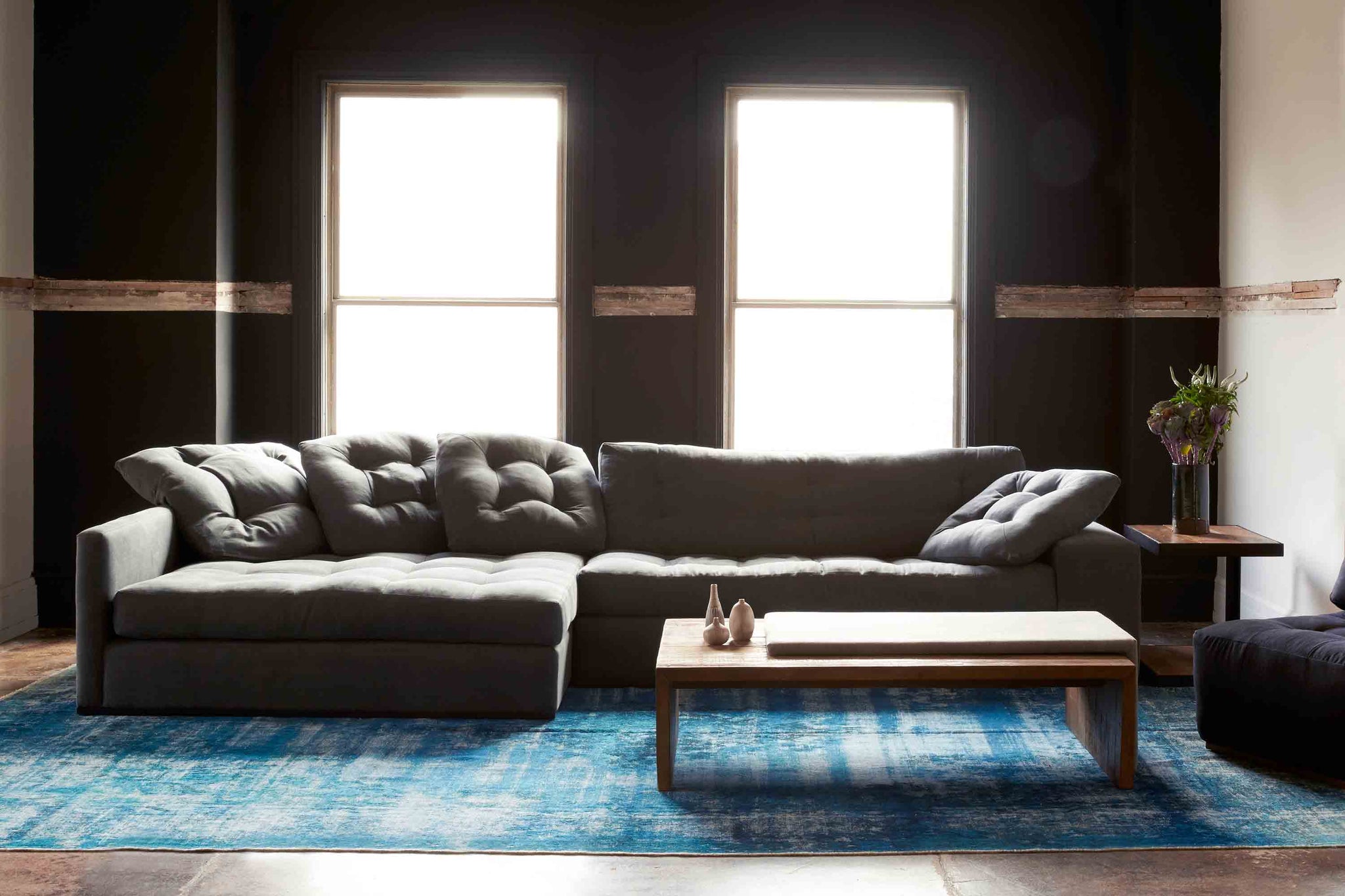  Studio Sectional in Molino Slate next to a wood coffee table and dark ottoman. Underneath the sectional is a blue rug. In the background we see a dark colored wall light shining through the two windows. Photographed in Molino Slate. 