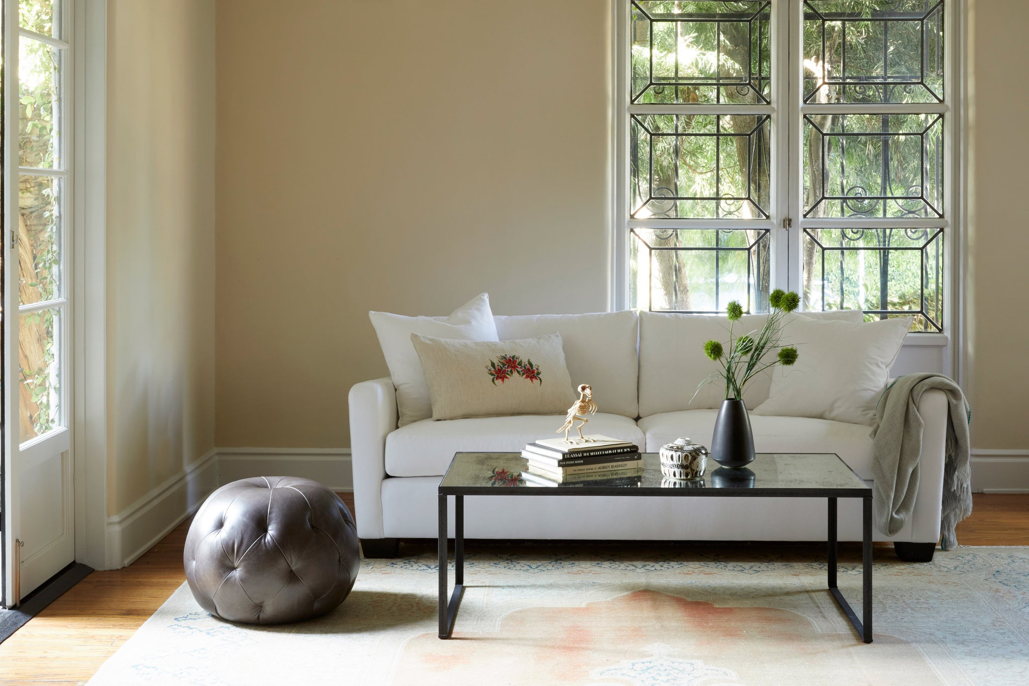  Sunset sofa upholstered in Denim White in a bright living room. A large window is behind the sofa and a leather pouf is on the side. A rectangular coffee table is in front with books and flowers on top.  Photographed in Denim White. 