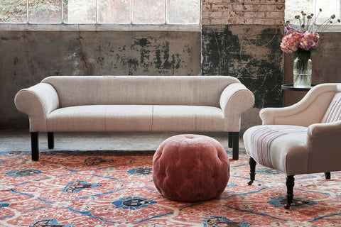 Teddy sofa in a light colored vintage fabric, with a chair on the right side, white fabric with some stripes. A pink velvet Pouf Ottoman is in front. In the background, there is a vase with flowers on a side table. Photographed in Vintage Hemp.