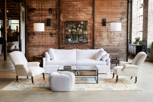  Tonto Ottoman in Pierre Stone next to a white sofa, white chairs, and a glass coffee table. Photographed in Pierre Stone. 