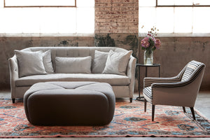  Large square ottoman in front of a grey sofa with a stripped chair on the right. Photographed in Lan Espresso. 
