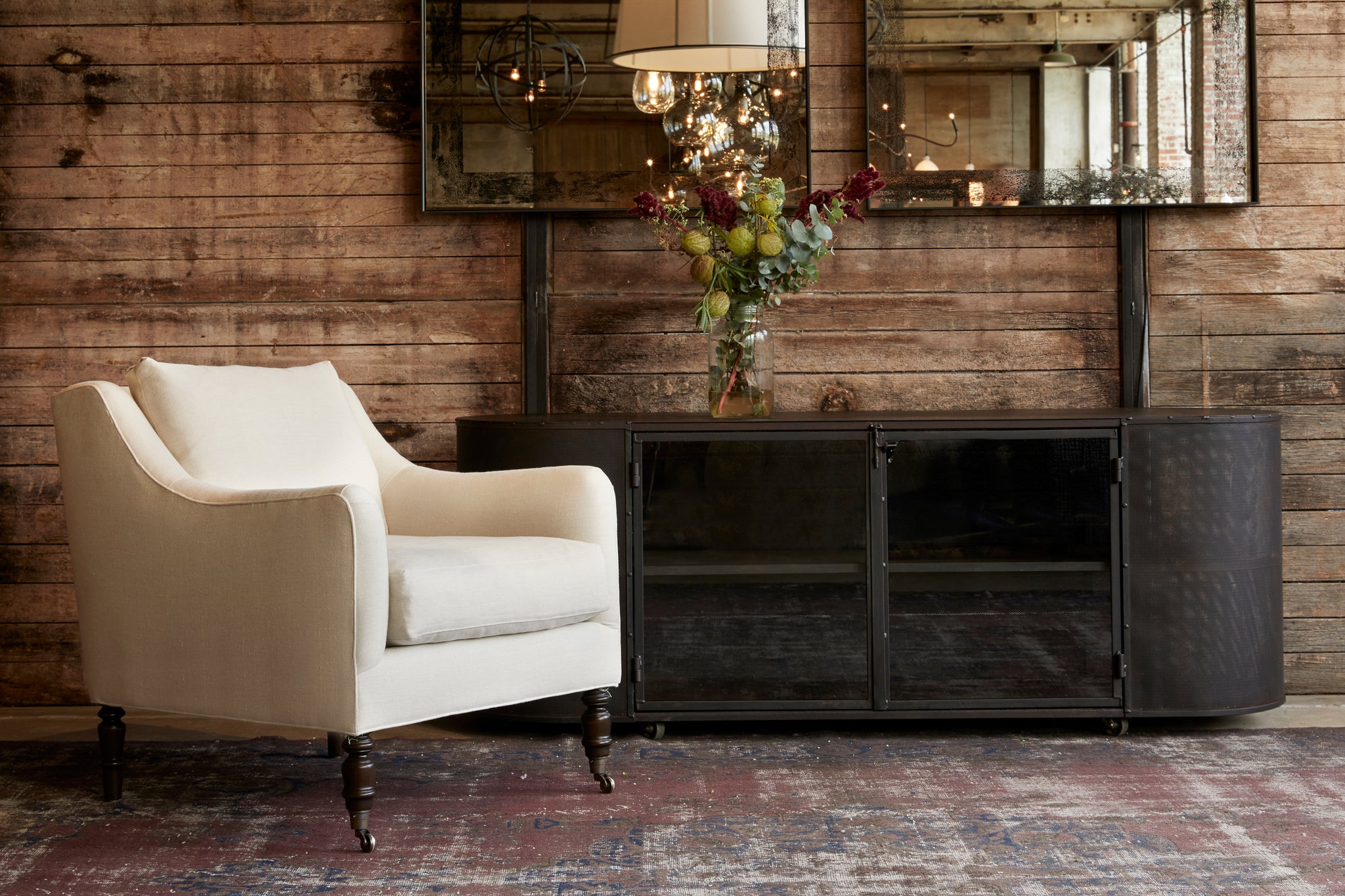  Yvonne chair in Noah Bone next to a dark credenza. The chair sits on a vintage rug. In the background is a wood wall with a metal mirror hanging. Photographed in Noah Bone. 